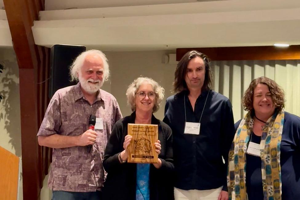 Four people standing side-by-side smiling. The third person from the left, a woman, is holding a wooden award plaque.